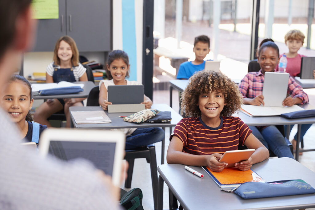 Smiling elementary school students sitting at desks and holding tablets 