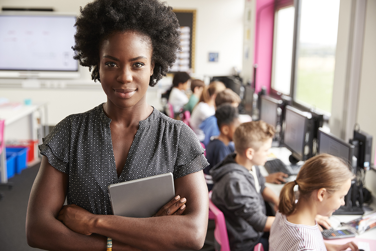 Teacher holding a tablet in her crossed arms stands in front of row of students working on computers